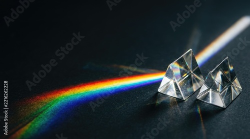 Three clear triangular prisms refract a vibrant rainbow light beam onto a dark surface.