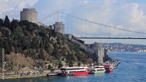 Suspension bridge spans wide waterway. Ancient stone fortress rises beside hillside. Several boats dock near waterfront promenade. Trees cover slopes under clear sky in Istanbul, Turkey