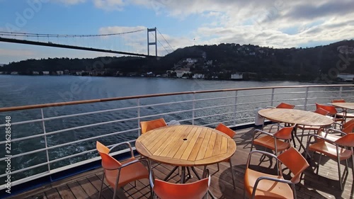 Boat deck with tables, chairs, distant bridge. Ocean and hills form scenic backdrop. Clouds drift above waters. Sunlight glows on wooden surfaces. Viewers enjoy waterfront ambiance in Istanbul, Turkey