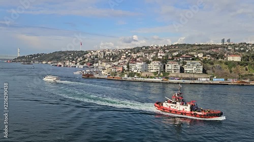 Red boat navigates busy waterway. Cityscape lines distant shoreline. Sky shows scattered clouds above. Boats move through calm blue waters. Water ripples trail behind vessels in Istanbul, Turkey