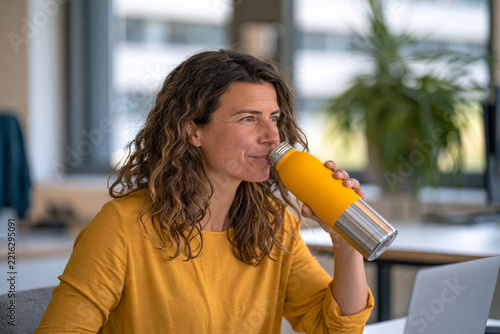 Young professional drinking from a reusable stainless steel water bottle while working at a modern minimalist desk, clean workspace, natural daylight