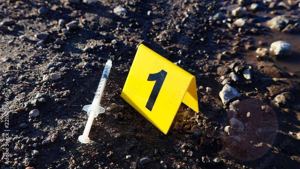 © Dark Artistic - Crime scene marker number one identifying a used syringe lying on a dark, wet, and rocky muddy ground, representing drug abuse, investigation, and legal concepts © Dark Artistic - Crime scene marker number one identifying a used syringe lying on a dark, wet, and rocky muddy ground, representing drug abuse, investigation, and legal concepts