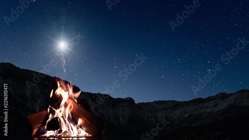 A campfire flickers beneath a brilliant star and a starry sky above dark mountains, evoking Christmas Eve.