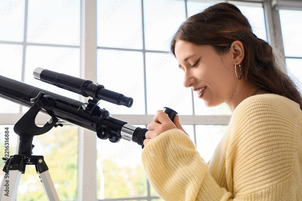 Fototapeta premium Stem education and scientific discovery, young woman looking through a telescope to explore astronomy and space science in a bright home interior