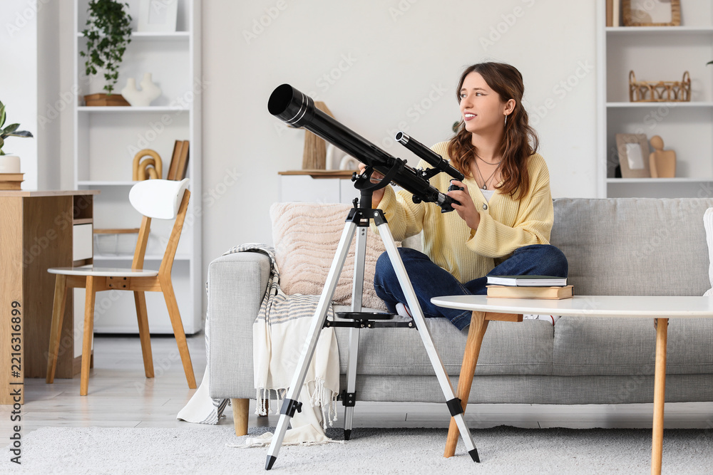 Fototapeta premium Astronomy and curiosity, young woman using a telescope at home for stargazing and science education in a cozy living room interior with modern decor