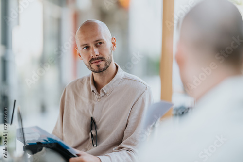 A bald man listens attentively during a business meeting while holding papers. The colleague speaks across the table in a casual office cafe conversation.