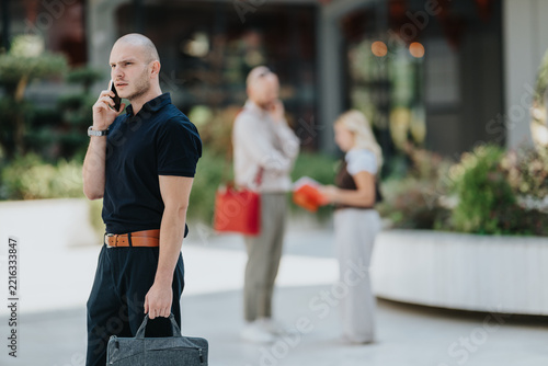 A businessman stands outdoors talking on his smartphone while holding a briefcase. Two blurred colleagues speak in the background, suggesting a professional urban setting.