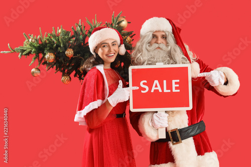 Young couple in Santa costumes with Christmas tree pointing at sale sign on r...