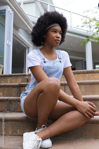 African American woman sitting on stone steps at modern porch wearing denim overalls and headband