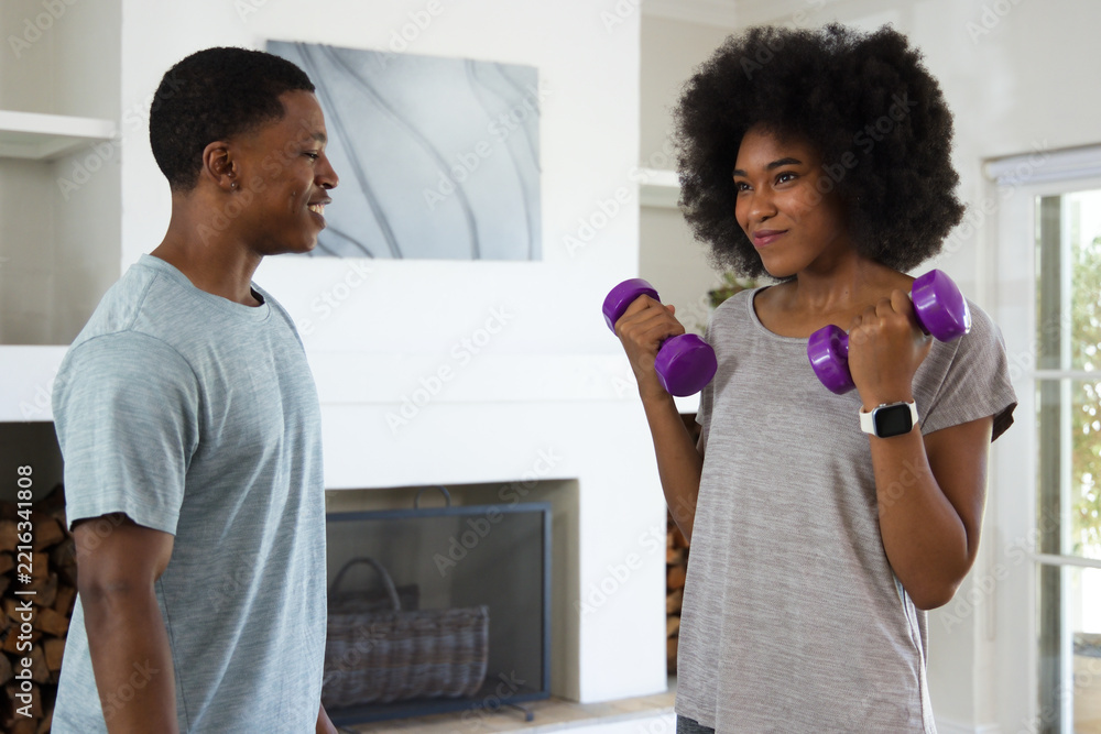 Naklejka premium African American couple exercising in living room, woman holding purple dumbbells and tracker