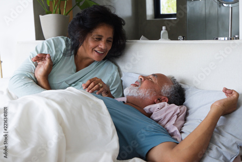 Senior couple smiling while leaning on padded headboard in bedroom with white duvet, ring visible