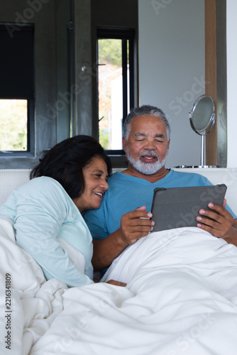 Senior African American couple sitting in bed under white duvet in modern bedroom, holding tablet