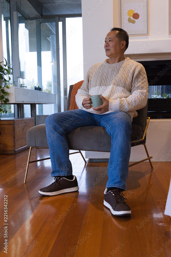 Fototapeta premium Senior Asian man sitting on grey chair at home wearing cream sweater holding light green mug