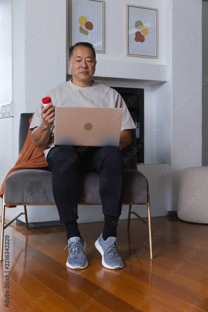 Fototapeta premium Senior Asian man sitting in living room on gray chair using laptop and holding red-capped bottle