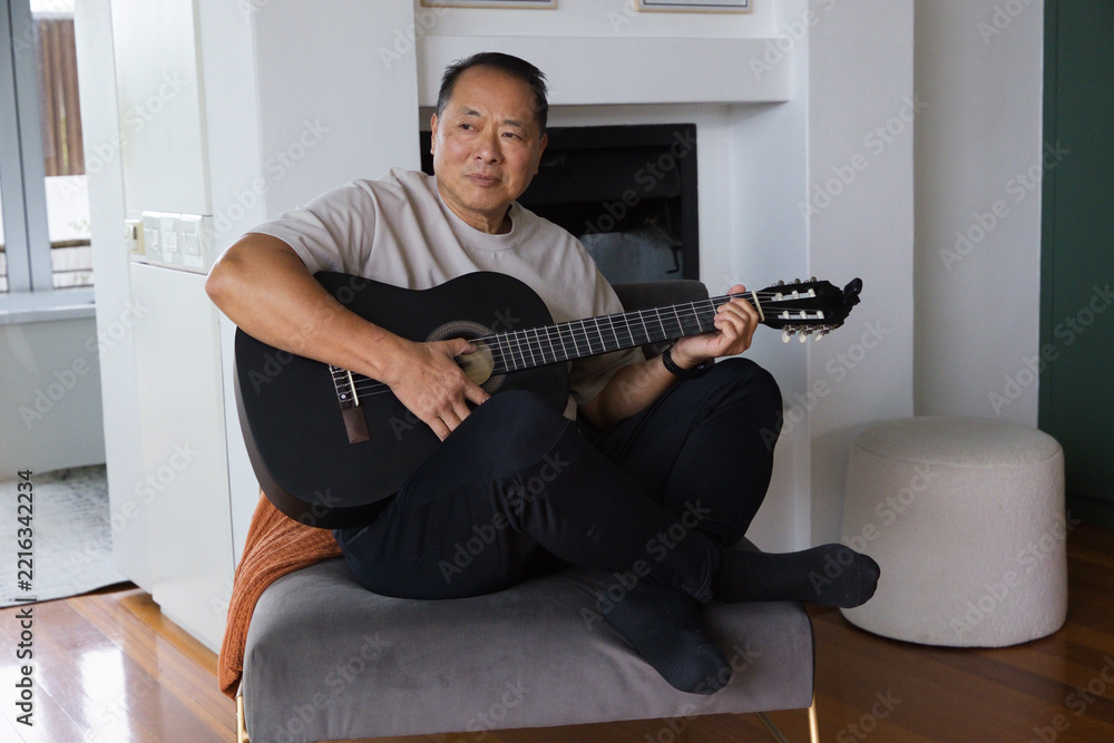 Fototapeta premium Senior Asian man sitting cross-legged on bench in living room by window holding black guitar