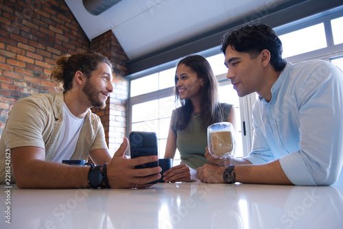 Diverse friends leaning at glossy white table in modern lounge, holding smartphone and glass mug