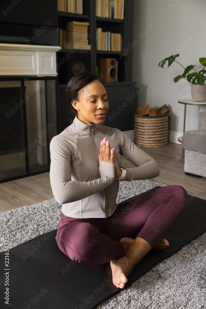Fototapeta premium Adult woman sitting cross-legged at home on black yoga mat, wearing gray jacket, leggings and watch