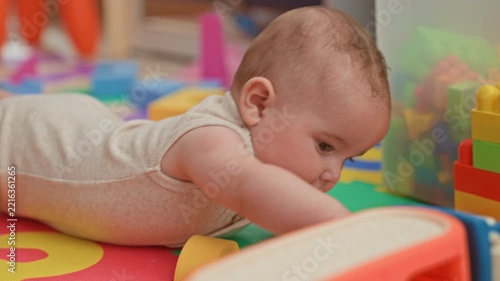 Baby on tummy reaching for a colorful toy on a foam playmat, hand gripping a block in building; curiosity innocence.