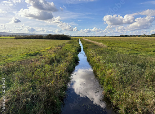 Rural countryside scene with green fields and blue sky with white clouds reflected in a stream stretching into the distance
