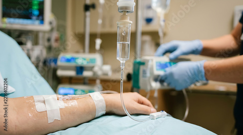 Close up of a patient receiving intravenous fluid through an IV drip in a hospital room with a nurse adjusting equipment.