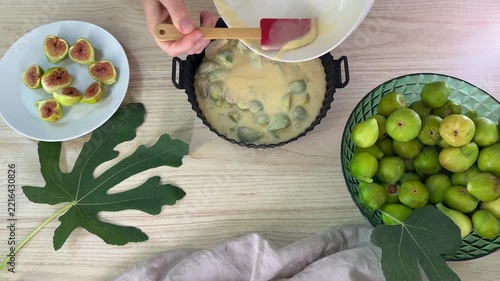 A person pours a pale yellow batter from a white bowl into a black fluted baking pan, surrounded by green figs and sliced fig pieces on a wooden surface