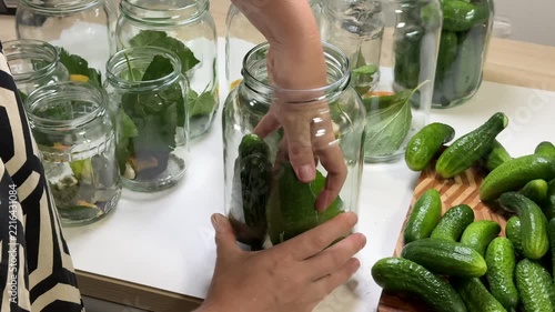 a person puts small green cucumbers into a clear glass jar, surrounded by other jars with green leaves and more cucumbers on a wooden board