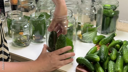 a person places a green cucumber into a glass jar among other jars filled with cucumbers and green leaves on a white table