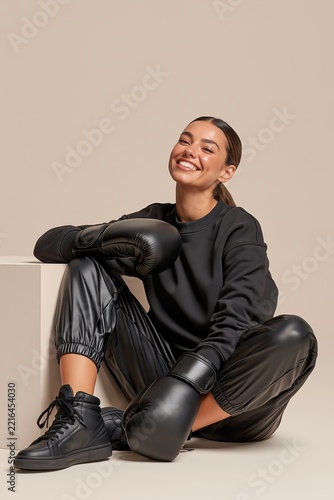Young woman in boxing gloves sitting on studio floor smiling confidently in black athleisure outfit.