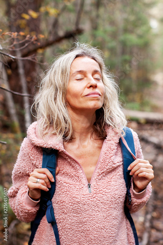 Middle aged woman pausing on a woodland walk with backpack, eyes closed. Digital detox and mindful time outdoors. Hiking, nature connection and screen free wellbeing. Active lifestyle