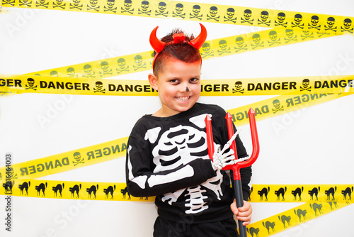 A portrait of a young smiling boy in a creative Halloween costume combining a skeleton-printed sweatshirt, red devil horns on a headband, and simple face paint. 