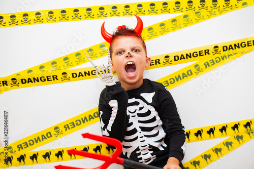 Excited little boy in a spooky Halloween skeleton costume and red devil horns making a rock hand gesture, standing against a white background decorated with yellow danger caution tape.