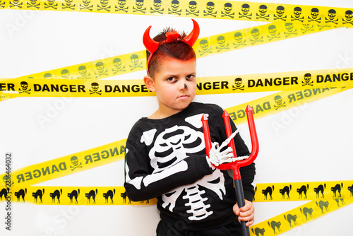 A portrait of a young upset boy in a creative Halloween costume combining a skeleton-printed sweatshirt, red devil horns on a headband, and simple face paint. 