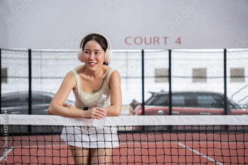 Fotografie Asian woman wearing headphones holding water bottle and leaning on net taking a