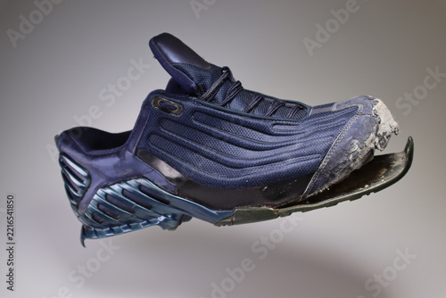 Single heavily worn blue athletic sneaker with torn detached rubber sole floating in air over grey studio background. Broken shoes concept.