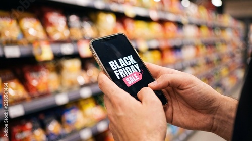 Man holding smartphone with Black Friday Sale on screen in supermarket aisle, grocery shopping, mobile shopping, discount, deal, close-up hands