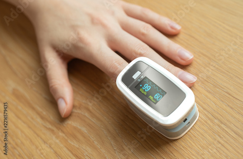 Pulse oximeter sensor clipped to fingertip reads oxygen saturation at 99% and pulse rate 60. Portable SpO2 device monitors vital signs as hand rests on wooden table. Compact gadget for home checkup.