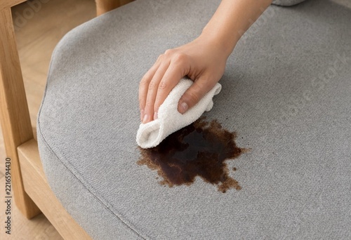 Close up of person wiping a dark liquid coffee stain off a gray upholstered chair with a white cleaning cloth