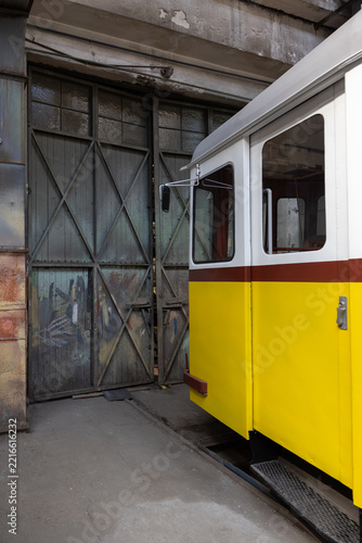 Vintage yellow and white tram car parked in front of an old industrial factory workshop sliding door, transportation heritage.