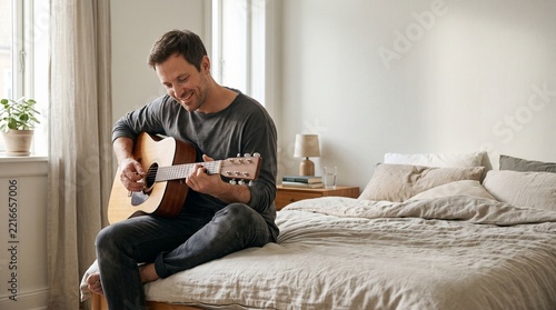 Homem tocando violão no quarto com expressão feliz, aproveitando momento de lazer e música em casa em fotografia natural para campanhas de estilo de vida