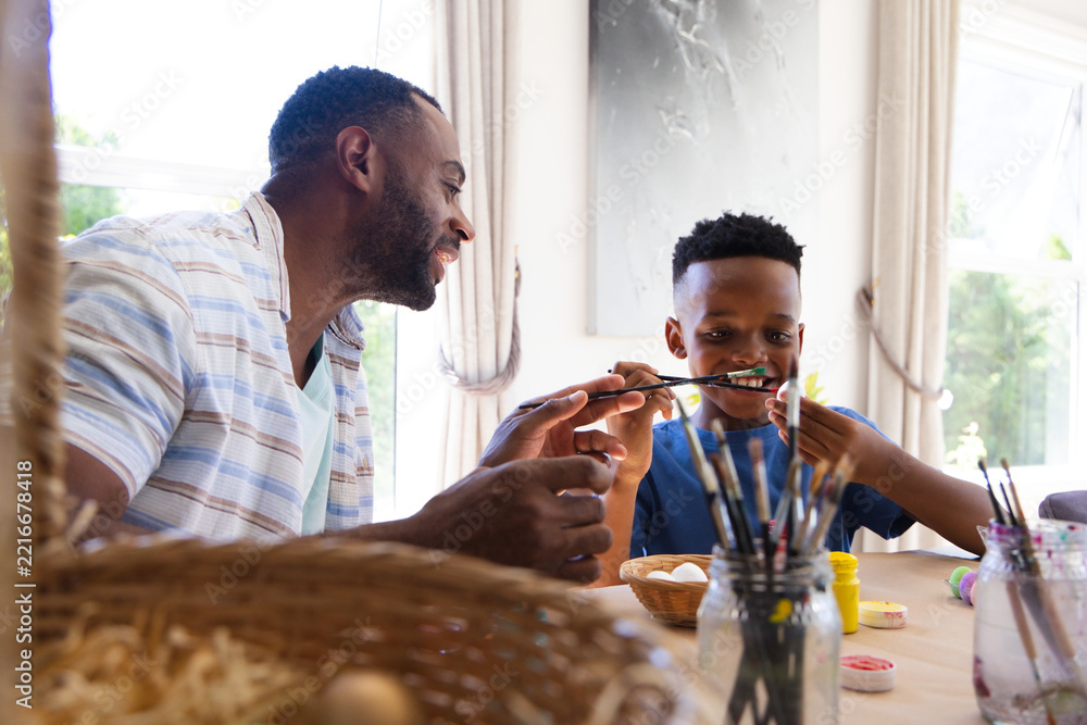 Naklejka premium African American father and youth son painting eggs at dining table by windows with wicker basket