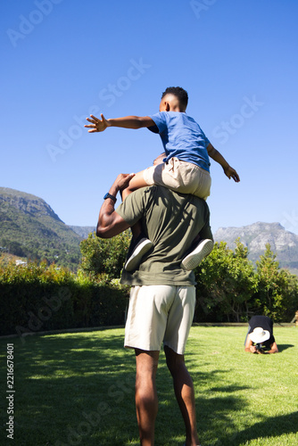 Mature African American father carrying youth son on shoulders in park lawn, showing wristwatch