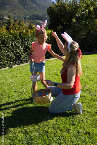 Mom and daughter kneeling in garden wearing bunny ears with baskets and eggs mom's smartwatch