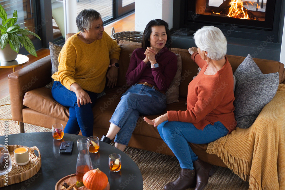 Fototapeta premium Diverse female friends sitting on brown leather sofa in living room, sipping drinks by fireplace