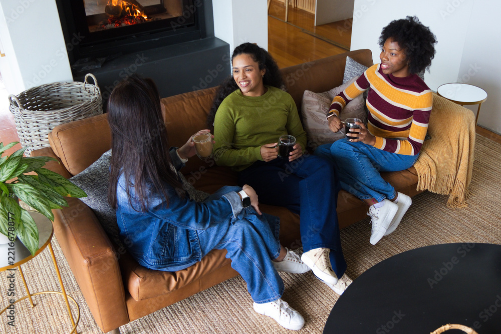 Fototapeta premium Diverse female friends chatting on leather sofa in living room beside lit fireplace holding mugs