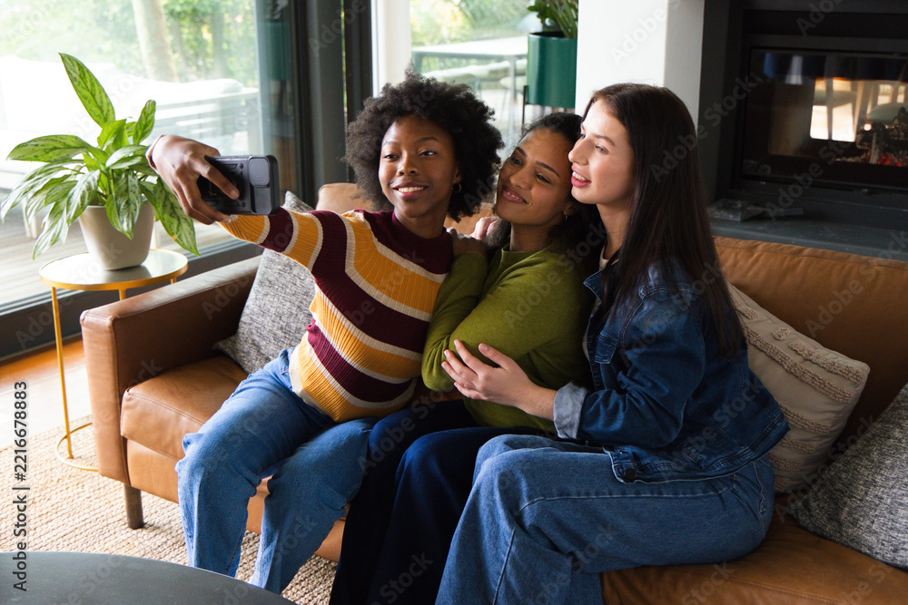Fototapeta premium Diverse female friends posing for selfie on tan leather sofa in living room with smartphone