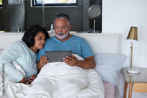 African American senior couple sitting upright in bed in modern bedroom, holding tablet and leaning
