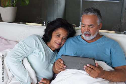 Senior couple reclining on bed in bedroom, holding tablet under white duvet wearing light blue tops