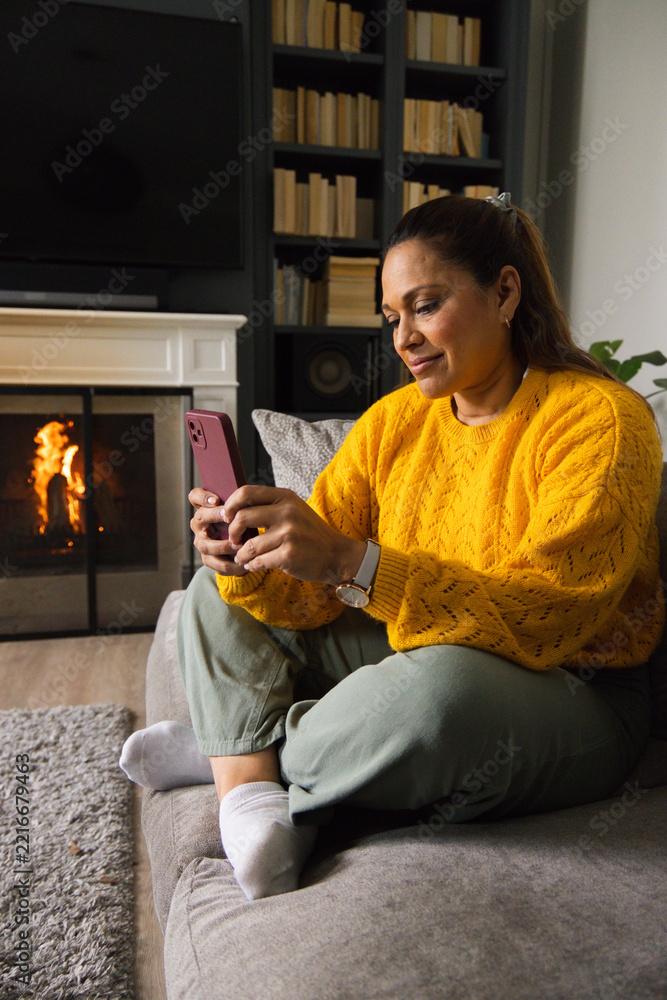 Fototapeta premium African American woman sitting on sofa wearing mustard sweater holding pink phone near fireplace