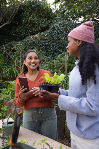 African American mother and teen daughter tending seedlings in garden holding red phone and trowel