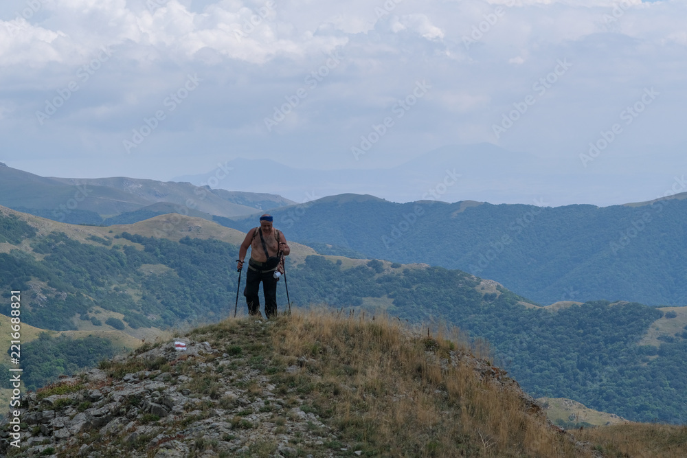 Obraz premium Overweight adult male traveler with a bare torso and large backpack walks alone through high green mountain peaks, moving heavily and persistently while carrying a load, representing endurance